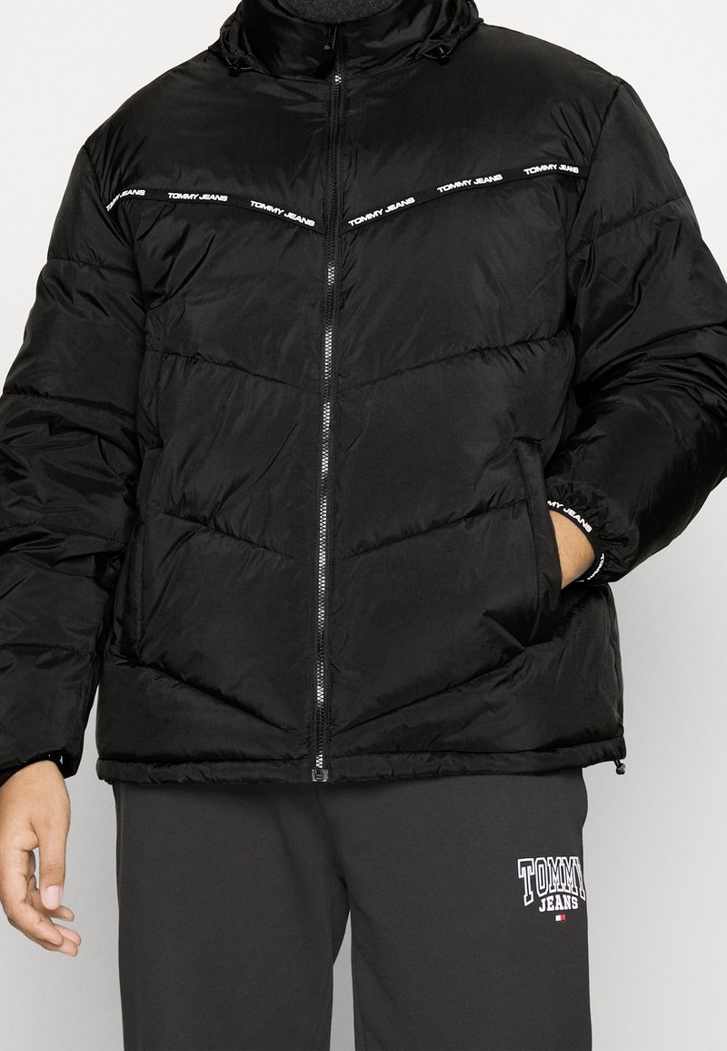Černá péřová bunda s vysokým límcem, zapínáním na zip a vodorovným prošitím. Na hrudi má bílé nápisy "TOMMY JEANS".