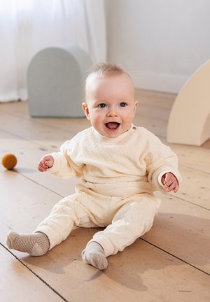 Tenue matelassée crème pour nourrissons avec manches longues et poignets élastiques, accompagnée de chaussettes gris clair et beige, assis sur un sol en bois.