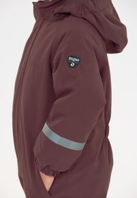 Bordeauxjack met een capuchon, gemaakt van zacht, waterdicht materiaal. Voorzien van een grijze reflecterende stripe en een zigzag badge op de mouw.