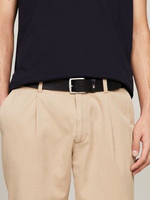 Schwarzer Leder-Gürtel mit silberner Metallschnalle, getragen mit hellbeigen Hosen und einem dunkelblauen ärmellosen Top mit sichtbaren Falten.