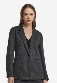 Marynarka w czarno-szary wzór w jodełkę z klapami, dwoma przednimi kieszeniami i zapięciem na jeden guzik. Teksturowany materiał.