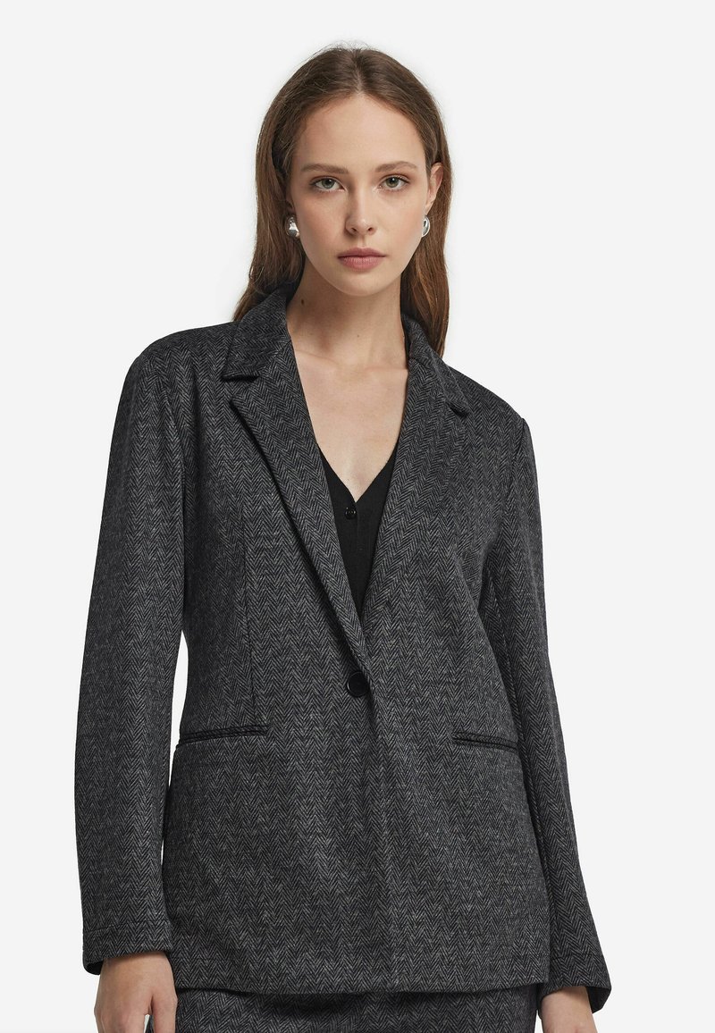 Marynarka w czarno-szary wzór w jodełkę z klapami, dwoma przednimi kieszeniami i zapięciem na jeden guzik. Teksturowany materiał.
