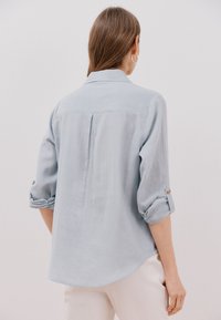 Camicia azzurro chiaro con bottoni, dotata di una piega sulla schiena, maniche arrotolate e un orlo curvo. Realizzata in un tessuto morbido con una consistenza liscia.