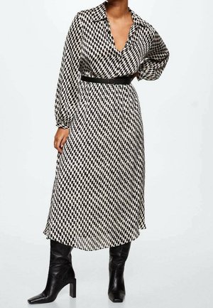 Schwarzes und weißes gemustertes Kleid mit kragenförmigem Ausschnitt, langen Ärmeln, tailliertem Bund und fließendem Rock, kombiniert mit knielangen schwarzen Stiefeln.