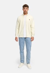 Hombre vestido con una chaqueta crema de cremallera sobre una camisa blanca, jeans azul claro y zapatillas crema, de pie contra un fondo liso.