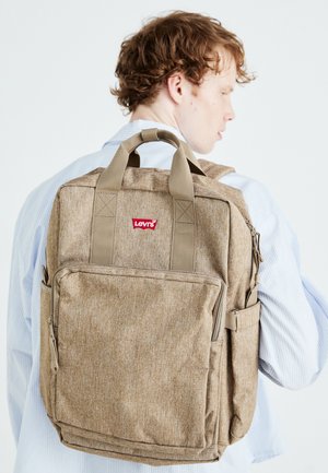 Brauner Stoffrucksack mit mehreren Taschen, stabilen Trägern und einem kleinen roten Logo. Verfügt über eine strukturierte Oberfläche und rechteckige Form.