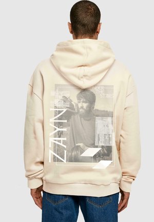 Beige hoodie lavet af blødt stof med et stort grafisk print på ryggen, med navnet "ZAYN" og yderligere designelementer.