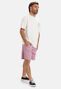 Man standing with hands in pockets wearing a white t-shirt, pink shorts, and black sneakers on a white background.