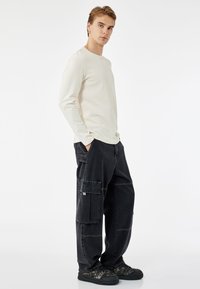 Krämfärgad långärmad skjorta med avslappnad passform, i kombination med svarta cargo-byxor med flera fickor och kontrasterande sömmar. Mörkt mönstrade sneakers.