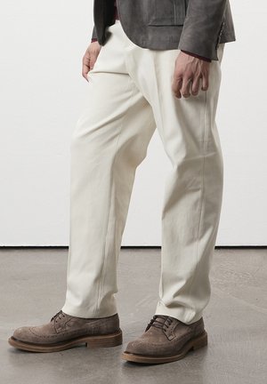 Un pantalon en coton de couleur crème présente une coupe décontractée avec des coutures latérales. Il est associé à des chaussures en daim marron et à une veste gris anthracite.