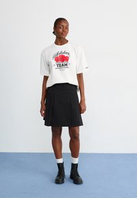 Hvid grafisk t-shirt med rødt blomsterdesign, parret med en sort plisseret nederdel og sorte snøreboots. Hvide ankelstrømper synlige.