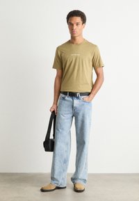 Olive green t-shirt with white text, light blue baggy jeans, tan shoes, and a black bag. Casual fit with minimalistic design elements.