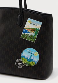 Ciemnobrązowa torba na ramię z czarnymi uchwytami, ozdobiona kolorowymi naklejkami podróżnymi Monako i Nowego Jorku oraz małym okrągłym logo stempla.