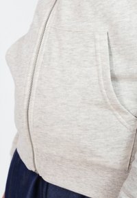 Sudadera de cremallera en gris claro, fabricada en una suave mezcla de algodón. Cuenta con un bolsillo frontal, una textura sutil y una cremallera visible en la parte delantera.