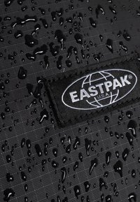 Materiale nero idrorepellente con gocce visibili. Presenta una toppa in tessuto nero con il logo "EASTPAK" bianco. Superficie strutturata.