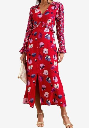 Femme portant une robe midi rouge fleurie à manches longues avec une fente devant, tenant une pochette dorée et portant des talons dorés à bride cheville.