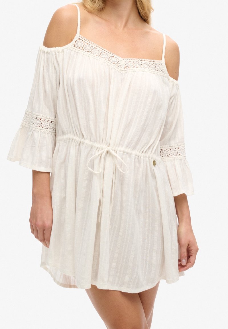 Robe crème à épaules dénudées avec des accents en dentelle, tissu léger et taille à cordon. Présente des manches évasées et des rayures verticales.