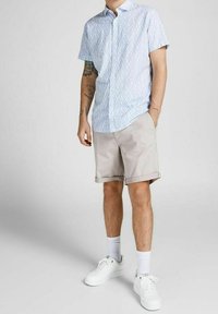 Světle modrá košile s krátkým rukávem a geometrickým vzorem, v kombinaci se světle béžovými šortkami a bílými teniskami. Viditelné hladké bílé ponožky.