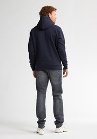 Námořnická modrá mikina s velkou kapucí, dlouhými rukávy a žebrovaným lemem, spárovaná se šedými džíny, zobrazená zezadu. Viditelné bílé tenisky.