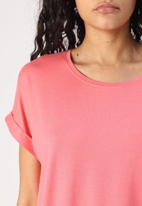 Rosa bomullströja med rundad halsringning, korta uppvikta ärmar och en slät, mjuk textur. Ingen mönstring eller ytterligare dekorationer.