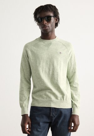 Lichtgroene trui met een gestructureerde breisel, ronde halslijn en lichte ribbels bij de mouwen. Gecombineerd met donkere jeans en een zwart zonnebril.