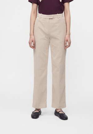 Person wearing beige straight-leg corduroy pants, dark purple top, and black Mary Jane style shoes standing against a plain light background.