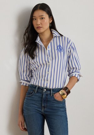 Pikkade tumedate juustega naine kannab valget särki siniste vertikaalsete triipudega, tikitud embleemiga, varrukad üles keeratud, siniseid teksapükse ja käevõrusid ühe randme ümber.