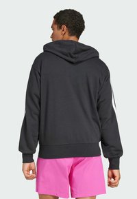 Sweatshirt preto com capuz, feito de um tecido suave, apresentando um bolso frontal tipo canguru, punhos canelados e riscas brancas nas laterais. Usado com calções cor-de-rosa.