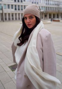 Bonnet tricoté beige clair, écharpe blanche crème et manteau rose clair avec des boutons bien en vue ; le mannequin pose à l'extérieur sur une surface carrelée.