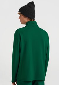 Langärmliges grünes Sportoberteil mit hohem Kragen, glatter Textur und Raglanärmeln, kombiniert mit einer schwarzen Strickmütze.