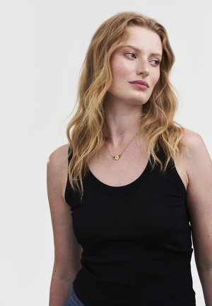 Woman with long wavy red hair in black sleeveless top looking sideways, wearing a gold necklace with a small flower pendant.