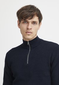 Pulover navy cu gât înalt, confecționat dintr-un material tricotat texturat, având un fermoar argintiu pentru închidere. Design simplu și modern.