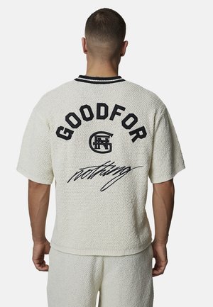 Homme de dos portant une chemise crème texturée et un short avec le texte noir "GOODFOR" en écriture cursive au dos de la chemise.