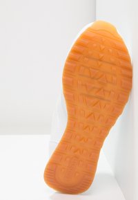 Bílá sportovní podrážka s texturovanou oranžovou gumou, s geometrickými vzory a logem. Boční profil na světlém pozadí.