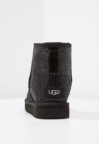 Černé ankle boty s texturovaným povrchem, zaoblenou špičkou a plochou podrážkou. Na boku mají logo UGG a jsou vyrobeny z měkkého textilního materiálu.