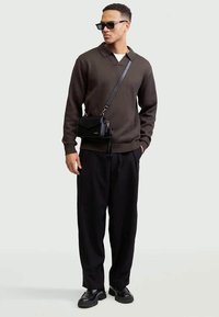 Uomo che indossa un maglione marrone scuro, pantaloni neri a gamba larga, mocassini neri, occhiali da sole neri e una borsa a tracolla in pelle nera.