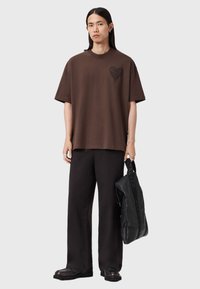 Brun bomuld T-shirt med et sort hjertegrafik på venstre bryst, kombineret med sorte vide bukser og en sort skuldertaske med struktur.