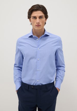 Jeune homme aux cheveux bruns ondulés portant une chemise à rayures bleu clair boutonnée et un pantalon bleu marine foncé, les mains dans les poches, fond neutre.