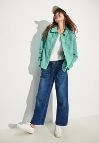 Veste en peluche de couleur sarcelle avec fermeture à bouton et deux poches sur la poitrine, associée à un pantalon en denim bleu ample et des baskets blanches. Casquette portée à l'arrière.