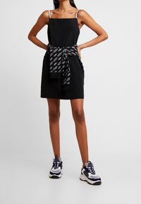 Frau in einem ärmellosen schwarzen Kleid mit weißen Logo-Trägern, einem schwarzen Sweatshirt mit Logo-Print um die Taille gebunden und navyblauen Sneakers mit weißen Akzenten.