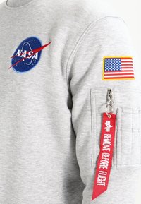 Felpa grigia con un patch del logo NASA e un patch della bandiera americana sulla manica. Include una tasca con zip con un tag di stoffa rosso etichettato "RIMUOVI PRIMA DEL VOLO".