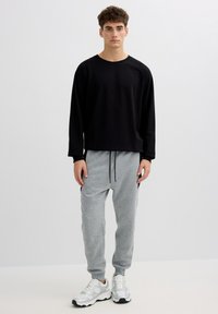 Hombre joven con una camisa negra de manga larga, pantalones deportivos grises con cordones negros y zapatillas blancas, posando frente a un fondo liso.