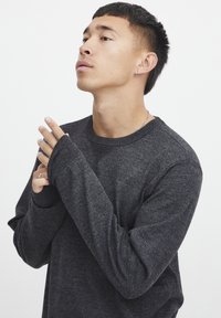Dunkelgrauer Pullover aus weichem Strickstoff. Mit Rundhalsausschnitt und langen Ärmeln mit Daumenlöchern. Das Modell posiert mit gefalteten Händen.