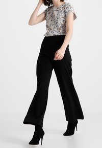 Camisa de estampado floral en gris con mangas arremangadas, combinada con pantalones negros de tiro alto con perneras acampanadas, llevados con tacones altos negros.