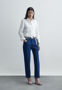 Λευκό πουκάμισο με μακριά μανίκια και κουμπιά, σκούρο μπλε τζιν παντελόνι tapered, ασημένια ζώνη με κούμπωμα και ροζ ψηλοτάκουνα παπούτσια. Απλό φόντο.