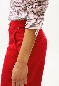 Pantalons rouges avec une texture lisse et une coupe ajustée, associés à une chemise blanche ornée de rayures verticales rouges et bleues.