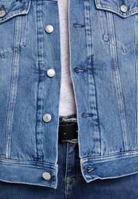 Giacca di denim blu chiaro con hardware argentato, dotata di chiusura a bottoni, due tasche sul petto e design con orlo grezzo. Indossata sopra una camicia bianca.