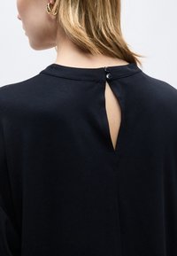 Γυναίκα με κοντά ξανθά μαλλιά φοράει μαύρο τοπ που κλείνει με δύο κουμπιά στο πίσω μέρος του λαιμού, αποκαλύπτοντας ένα μικρό άνοιγμα σε σχήμα κλειδιού.