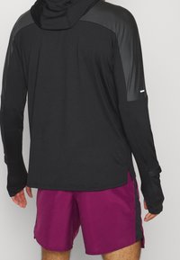 T-shirt de sport noir à manches longues avec capuche, présentant des panneaux texturés noirs aux épaules, associé à un short violet.