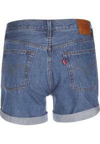 Kesäpikkupöksyt keskisinisessä farkkukankaassa, joiden helmassa on kulunut reuna. Varustettu kahdella takataskulla, joissa on keltaiset tikkaukset, ja vyötäröllä ruskea nahkainen brändilogo.